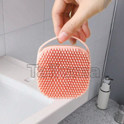 hand holding a silicone bath brush with soft bristles and a hanging strap, placed near a sink.